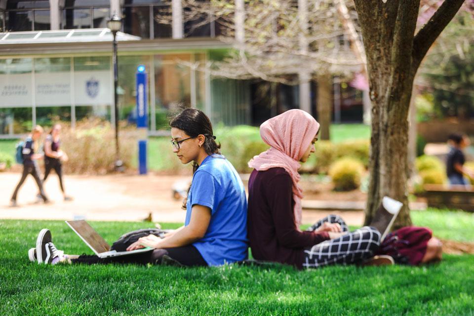 Two slu students sit back to back on the ground under a tree on the 博彩网址大全 Quad.